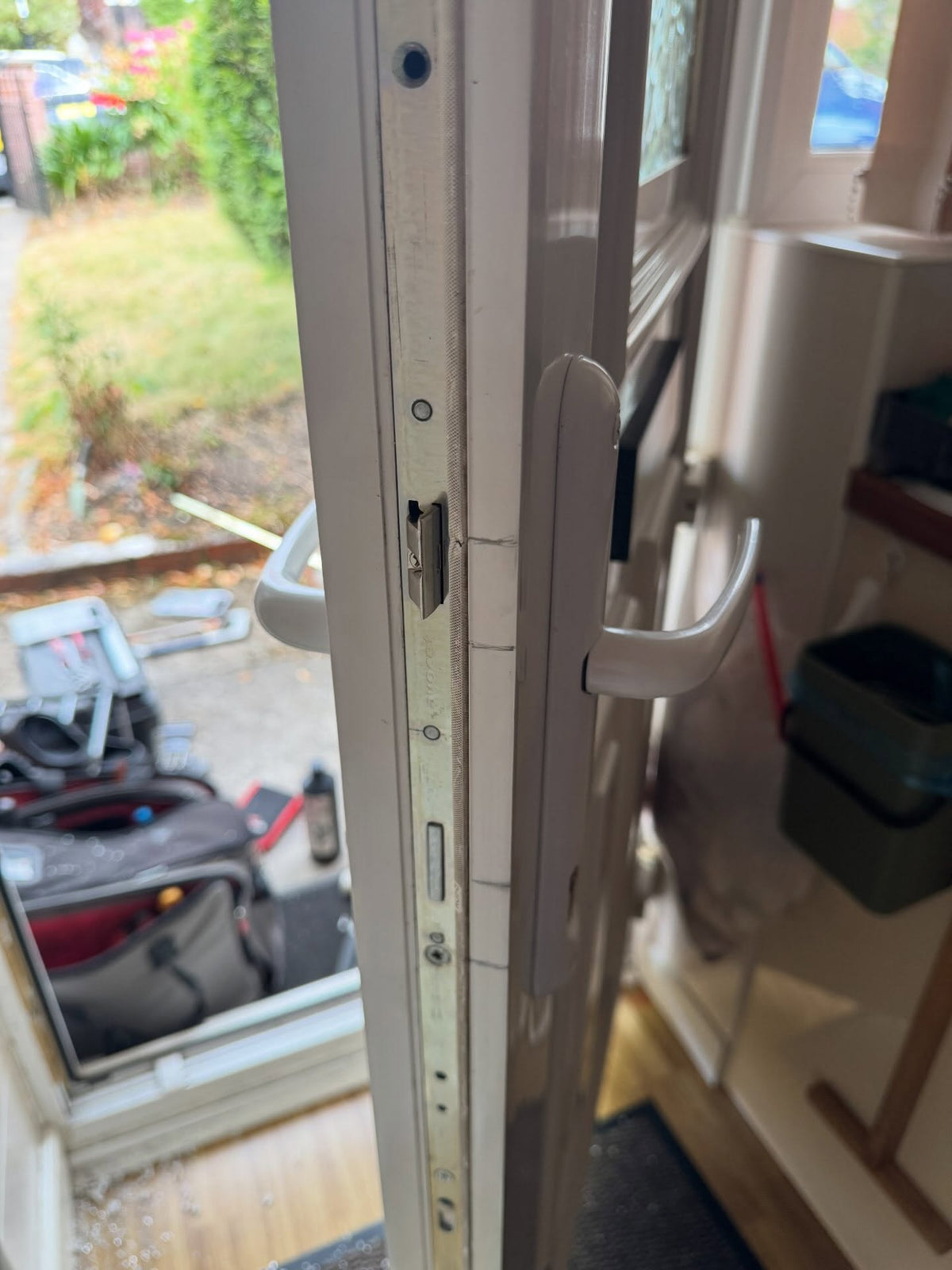 Close-up of a white composite door and handle set, showing the latch mechanism and door edge, with an open door and outdoor garden visible on the left and a home interior on the right.