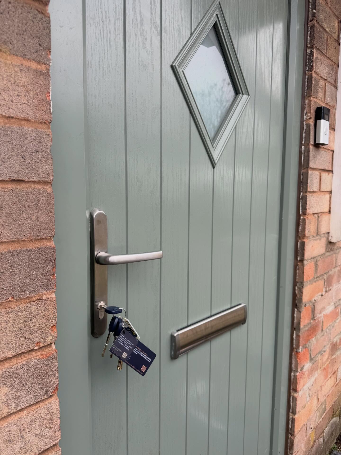 Gray front door with a diamond-shaped window and handle on a brick wall.
