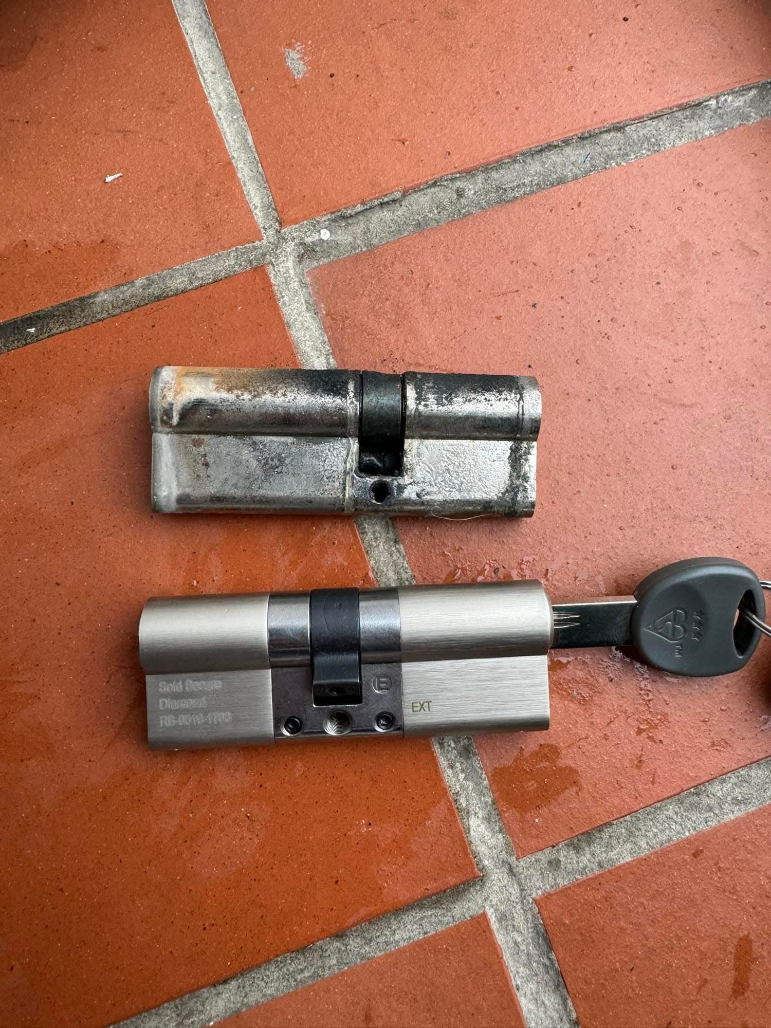Two metallic euro cylinder locks on a tiled floor with a key showing difference between basic euro lock and an anti snap 3 star lock